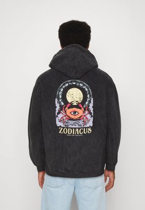 Osoba ubrana w czarną bluzę z kapturem z grafiką czerwonego kraba z okiem, pełnią księżyca i symbolami zodiaku, stojąca na jednolitym tle.