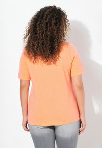 Camiseta de manga corta en algodón naranja brillante, con una textura suave y un dobladillo redondeado, combinada con unos vaqueros grises.
