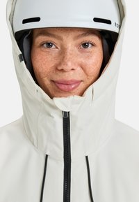 Weiße wetterfeste Jacke mit hohem Kragen, schwarzem Reißverschluss und Kordelzug, getragen unter einem weißen Helm. Sommersprossen und blaue Augen sichtbar.