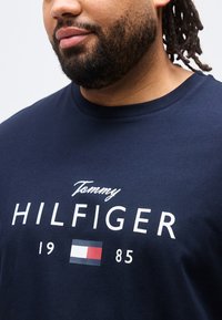 Άνδρας με γενειάδα και dreadlocks φορώντας μπλε σκούρο μπλουζάκι Tommy Hilfiger με λευκό λογότυπο και κόκκινη, λευκή και μπλε ορθογώνια σημαία.