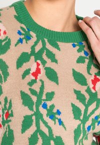 Pull en tricot beige avec des feuillages verts, des fleurs rouges et des accents bleus. Le col vert côtelé ajoute de la texture et du contraste au design.
