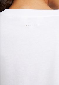 Camiseta de algodón blanca con cuello redondo y un sutil logotipo metálico plateado "Sapphire" ubicado en la parte trasera. Textura suave.
