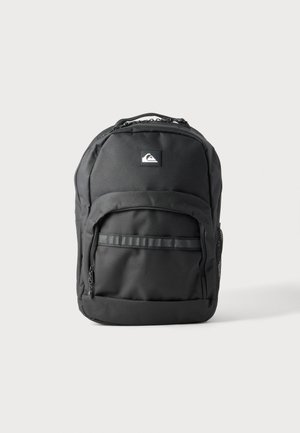 Sac à dos noir avec poche zippée à l'avant, poche latérale en mesh, poignée supérieure et logo Quiksilver sur le devant sur fond gris clair.