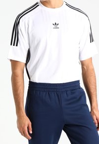 White athletic t-shirt with black three-stripe design on sleeves and small black Adidas logo. Paired with navy blue elastic waistband pants.