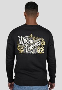 Svart sweatshirt med ett dekorativt ryggdesign som har orden "Fel Vänner" i vit och guldig utsmyckad text. Standard passform.