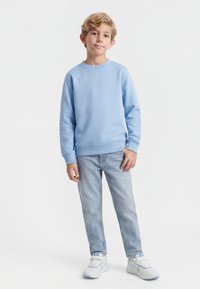 Jonge jongen die staat, gekleed in een lichtblauwe sweatvest, lichtblauwe spijkerbroek en witte sneakers, tegen een effen witte achtergrond.