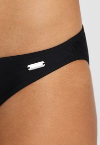 Schwarze Bikini-Hose in einem eleganten Design, mit einem silbernen Hardware-Tag mit dem Text "Venice Beach". Glatte Textur, minimalistische Form.