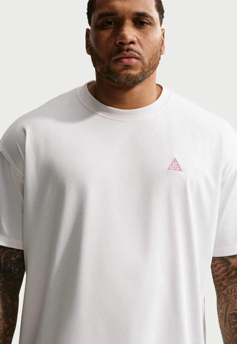 Homme aux cheveux courts et tatouages sur les bras, portant un t-shirt blanc uni avec un petit logo en forme de triangle rose sur la poitrine.