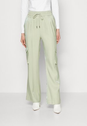 Femme portant un pantalon cargo large vert clair avec taille à cordon et grandes poches latérales, associé à des bottes blanches et un haut à manches longues.