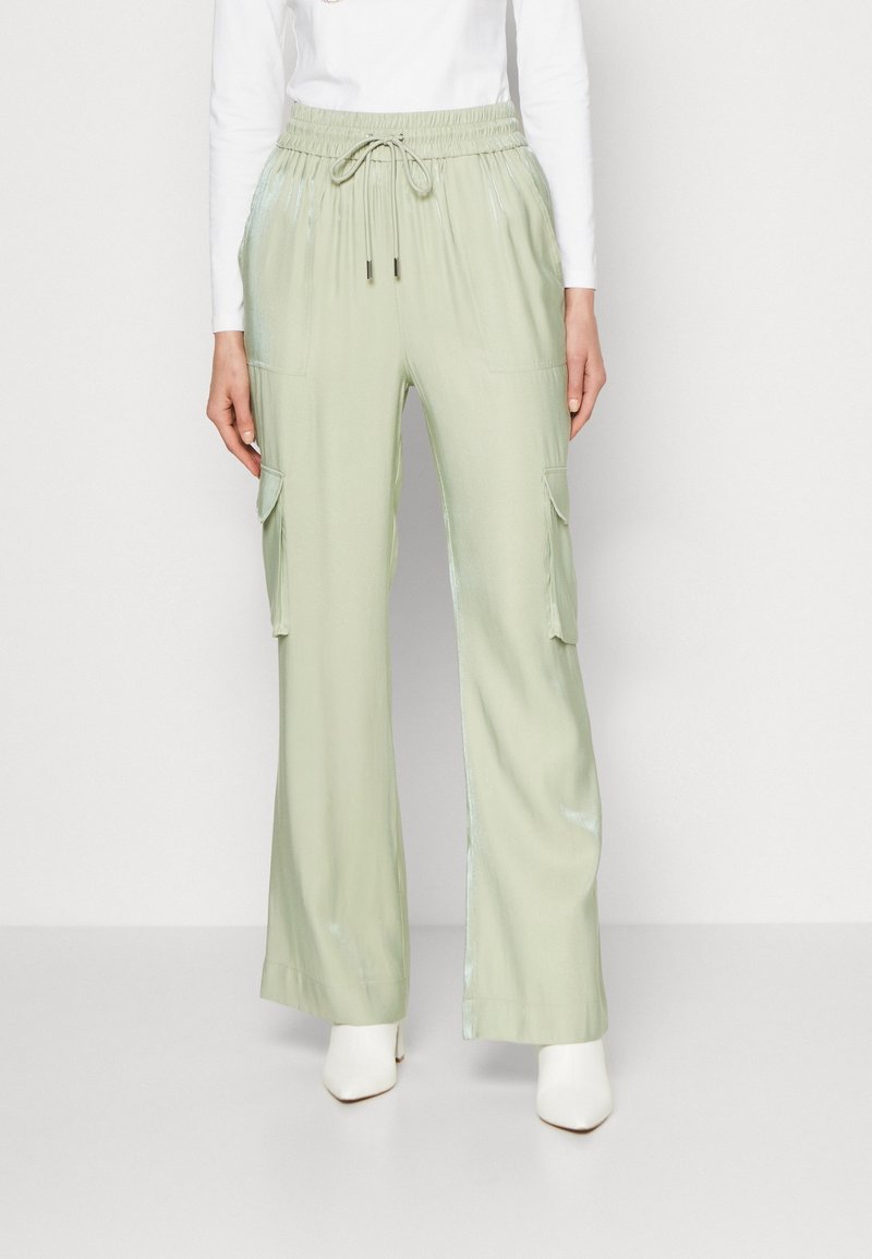 Femme portant un pantalon cargo large vert clair avec taille à cordon et grandes poches latérales, associé à des bottes blanches et un haut à manches longues.