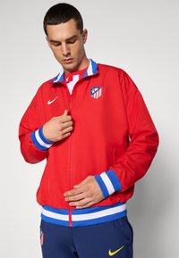 Red sports jacket with blue and white striped collar and cuffs, featuring a logo on the chest and a zippered front. Smooth material, relaxed fit.