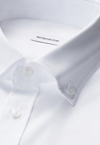 Zbliżenie na biały kołnierz i listwę z guzikami koszuli zapinanej na guziki, z widocznymi guzikami oraz metką marki "seidensticker" wewnątrz kołnierza.