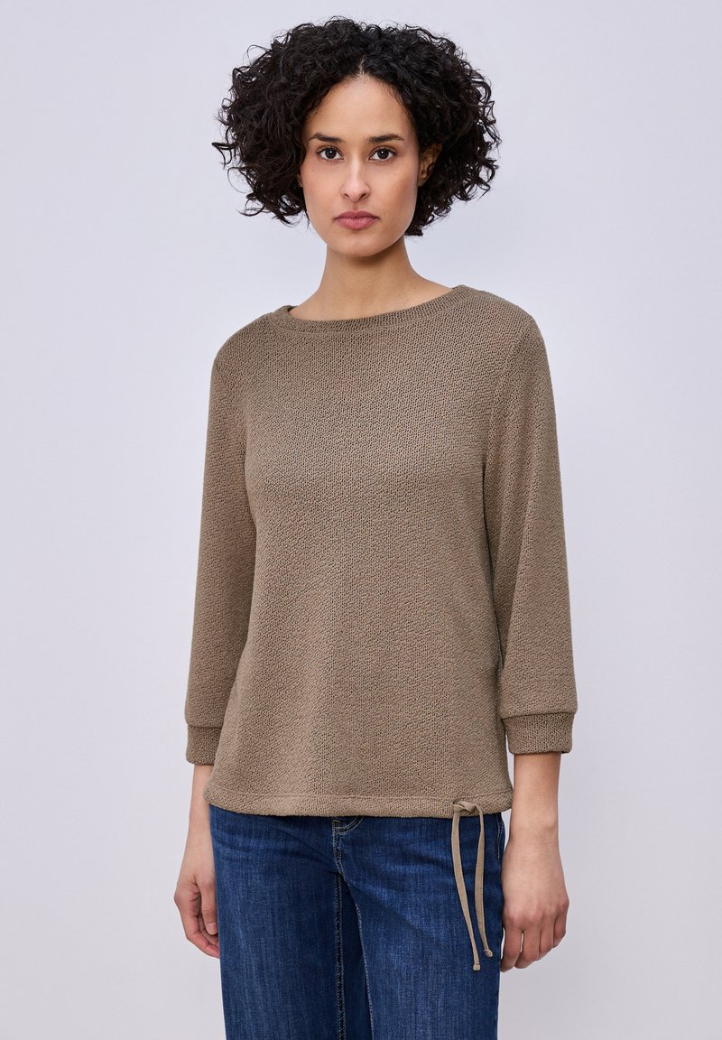 Femme aux cheveux bouclés portant un pull texturé taupe à manches trois-quarts et un jean bleu, sur un fond clair uni.