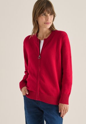 Rød zip-up cardigan med ribbet krave og manchetter, der har en blød tekstur og en afslappet pasform, båret over en hvid top og blå bukser.