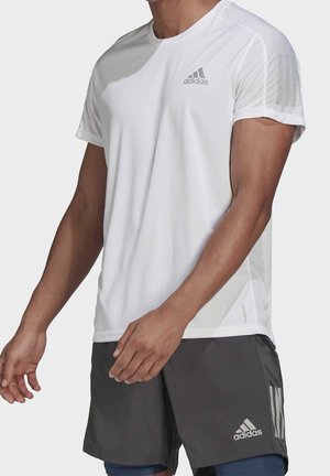 Homme portant un t-shirt de sport blanc à manches courtes Adidas et un short Adidas gris foncé sur fond uni.
