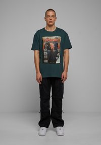 Mørkegrøn oversized T-shirt med grafisk print, parret med sorte cargo-bukser med lommer. Model iført hvide sneakers.