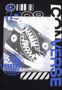 Camiseta negra con un gráfico de zapatos Converse de caña alta negros, con cordones blancos y un logo estrellado, complementada por texto azul y elementos de diseño.