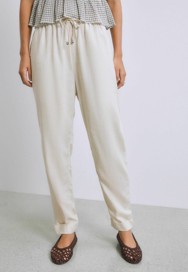 Femme portant un pantalon beige ample avec cordon de serrage, associé à des chaussures plates tressées marron, debout devant un fond clair uni.