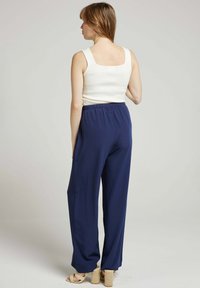 Débardeur blanc à bords festonnés, associé à un pantalon large bleu marine ; modèle portant des sandales compensées beiges, vu de dos.