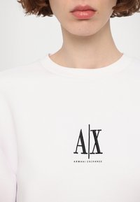 Biely sveter s logom "A|X" v čiernej výšivke, s textom "ARMANI EXCHANGE" nižšie. Mäkka textúra a dizajn s okrúhlym výstrihom.