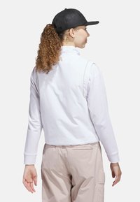 Hvid langærmet top med høj krave, der har en glat tekstur, parret med lyse beige bukser. En sort kasket fuldender outfittet.