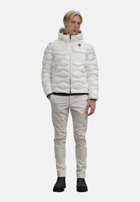 Giacca a vento bianca con design trapuntato e texture, chiusura frontale con zip e cappuccio. Abbinata a pantaloni beige chiaro e stivaletti neri.