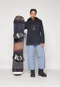 Veste imperméable noire avec capuche, pantalon de neige bleu et bottes noires, tenant un snowboard avec un dégradé de brun foncé et clair.