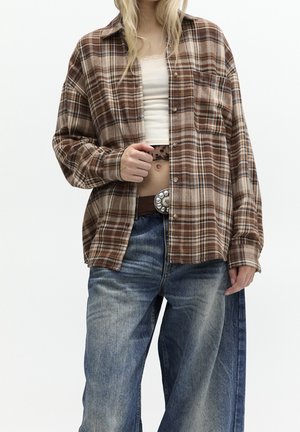 Personne portant une chemise à carreaux marron boutonnée par-dessus un haut blanc court, un jean bleu et une ceinture avec une boucle décorative, tenant la chemise ouverte.