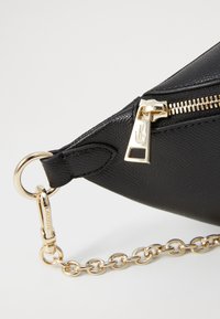 Borsa in pelle nera con finitura testurizzata, hardware dorato, tracolla a catena e chiusura con zip caratterizzata da un dettaglio logo distintivo.