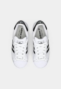 White athletic shoes with black stripes, rubber toe cap, and flat laces. Features an embossed logo on the tongue and cushioned heel.