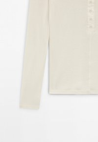 Chemise henley beige à manches longues avec quatre boutons sur le plastron, vue de la moitié du corps jusqu'au poignet sur un fond blanc.