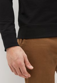 Svart långärmad tröja med rundad halsringning, tillsammans med bruna byxor. Närbild av en hand som vilar på byxbenet, vilket visar textur och passform.