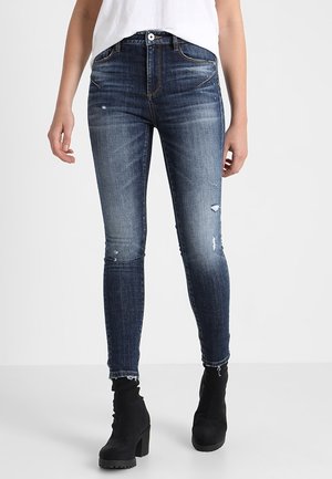 Woman wearing dark blue distressed skinny jeans with frayed hems and black heeled ankle boots, standing against a plain white background.