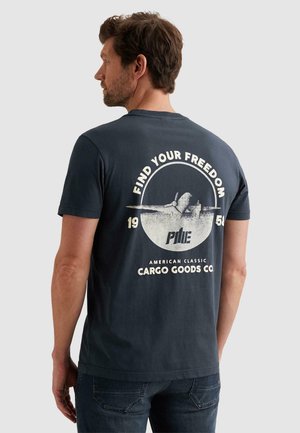 Mann trägt ein dunkelblaues T-Shirt mit einem Vintage-Flugzeug-Motiv und dem Text "Find Your Freedom, 1950, Cargo Goods Co." auf der Rückseite.