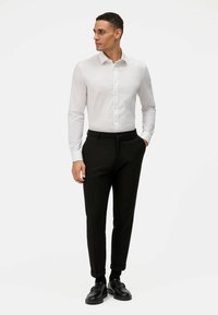 Weißes Langarmhemd mit Knöpfen, kombiniert mit schwarzen Tailor-Hosen und schwarzen Schuhen. Glatter Stoff, schlanker Schnitt, aufgerollte Säume an der Hose.