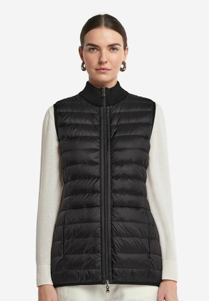 Schwarze Steppweste mit hohem Kragen, Reißverschluss vorne und gestepptem Muster. Über einem hellen Langarmpullover getragen, der eine taillierte Passform zeigt.