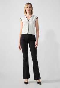 Hvit ermeløs bluse med svart kant og knapper, paret med svarte flared jeans og svarte hæler. Enkelt bakgrunn.