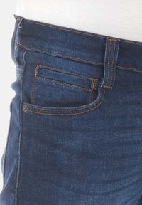 Jean en denim de couleur bleu foncé, avec des coutures orange, une poche frontale et un design de ceinture structurée. Texture lisse.