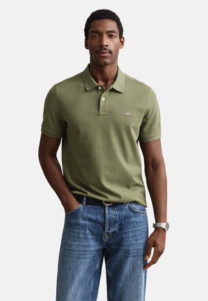 Hombre con camisa polo verde oliva de manga corta y vaqueros azules, de pie con una mano en el bolsillo frente a un fondo liso.