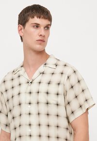 Camicia a maniche corte in un tessuto leggero e trasparente, con un motivo a quadri beige e nero, chiusura a bottoni e taschino sul petto.