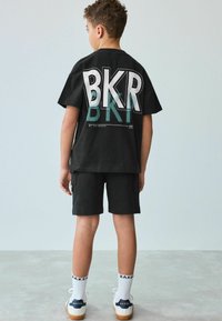 Čierne tričko s krátkym rukávom a veľkým bielym grafickým prevedením "BKR", kombinované s čiernymi šortkami a bielymi ponožkami so značkou "BAKER." Neformálny outfit s moderným dizajnom.