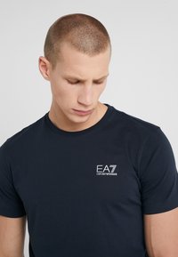 Námorně modré tričko s krátkým rukávem vyrobené z bavlny. Na levé straně hrudi se nachází malé bílé logo EA7. Klasický design s kulatým výstřihem.
