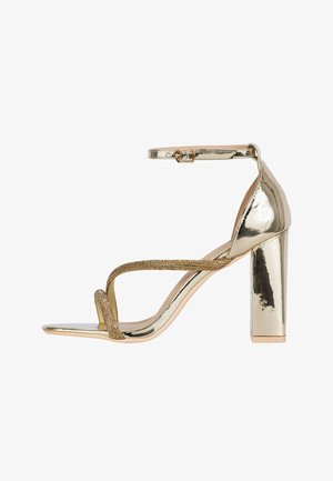 Zlaté metalické sandály na vysokém podpatku s páskem kolem kotníku, blokovým podpatkem, otevřenou špičkou a dvěma tenkými třpytivými předními pásky, které se kříží přes nohu.