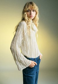 Blusa crema con texture, maniche lunghe e svasate, abbinata a jeans in denim blu. Tessuto morbido e leggero con pieghe verticali.