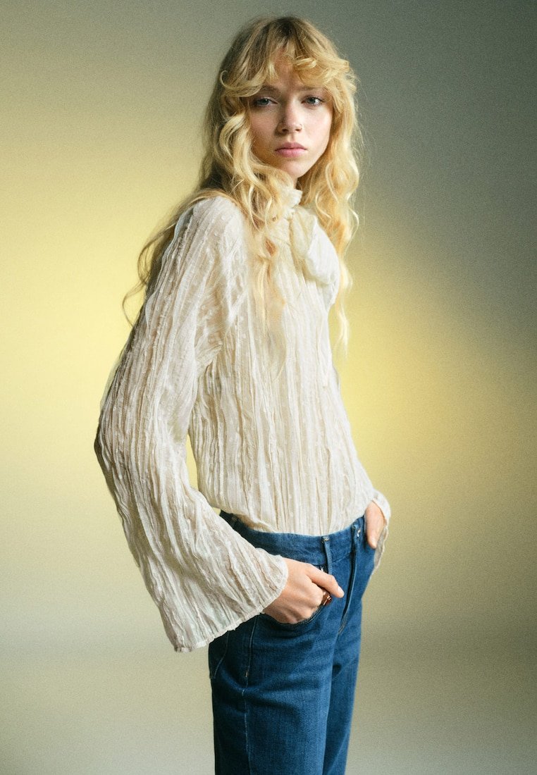 Blusa crema con texture, maniche lunghe e svasate, abbinata a jeans in denim blu. Tessuto morbido e leggero con pieghe verticali.