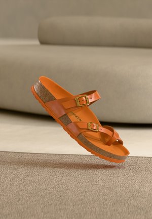 Sandale Birkenstock orange avec semelle en liège et deux brides ajustables avec boucles dorées, suspendue au-dessus d'un sol recouvert de moquette beige.