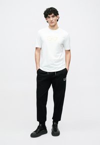 Λευκό κοντομάνικο t-shirt με χρυσό λογότυπο "EA7", σετ με μαύρα παντελόνια φόρμας που διαθέτουν διακριτικό λογότυπο και πλαϊνές τσέπες, συμπληρωμένα με μαύρα αθλητικά παπούτσια.