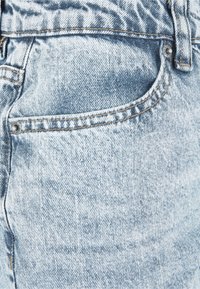 Primo piano della tasca di un paio di jeans in denim azzurro chiaro con cuciture visibili e rivetti metallici ai bordi.