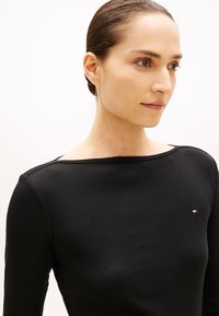 Crni top s dugim rukavima i izrezom u obliku broda, izrađen od mekanog materijala. Na lijevoj strani nalazi se mali logo u crvenoj, bijeloj i plavoj boji. Glatka tekstura.