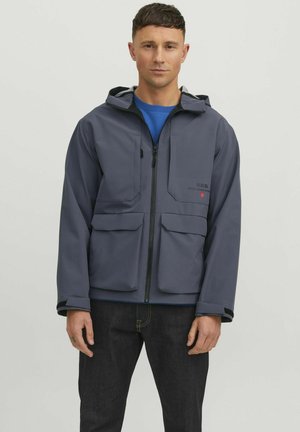 Homme portant une veste à capuche bleue avec des poches avant, une chemise bleue en dessous, et un jean foncé, se tenant devant un fond clair.